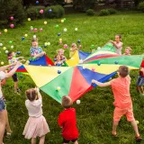 Niños y adultos juegan con un paracaídas de colores y pelotas, celebrando una fiesta de cumpleaños original en el césped.