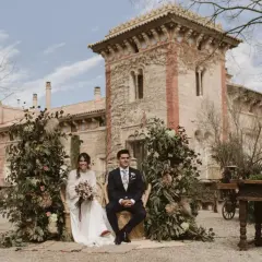 Novios sentados en sillas de mimbre, rodeados de exuberante vegetación y flores, con una arquitectura histórica de fondo. La decoración boda boho crea un ambiente romántico y natural.