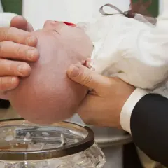 Manos sostienen a un bebé para el bautizo, con agua cayendo en una pila bautismal. Esto es parte de lo que se necesita para un bautizo.