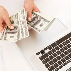 Manos contando billetes de 100 dólares sobre un portátil. Una forma de empezar a pensar en como ganar 100 euros al mes.