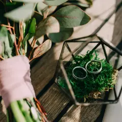 Anillos de boda en musgo verde dentro de un terrario de vidrio, junto a un ramo con tela rosa. Celebrando 5 años de casados.