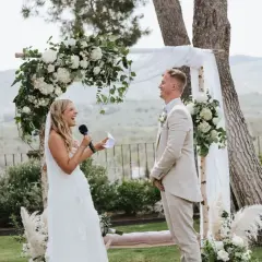 Novia ríe mientras lee sus votos, novio sonríe. Bodas sencillas pero bonitas, con arco floral y paisaje de fondo.