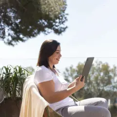 Mujer sonríe mientras trabaja en su tablet al aire libre, disfrutando de la vida de los trabajos para nómadas digitales.