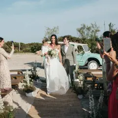 Novios caminan por la playa en una boda elegante. La novia lleva un niño en brazos. Un coche vintage espera.