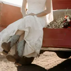 Novia con vestido de encaje y botas vaqueras, sentada en la parte trasera de una camioneta roja, con un ramo de flores. Un toque rústico para bodas vintage.