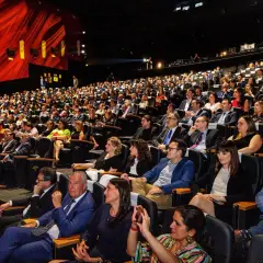 Amplia audiencia en un auditorio oscuro, asistiendo a eventos de empresa. El telón rojo brillante y las luces del escenario iluminan la sala llena de gente.