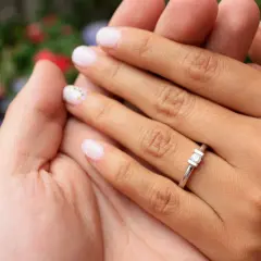 Manos entrelazadas, un dedo con un anillo de compromiso brilla entre flores.