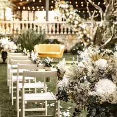Un arreglo floral blanco y dorado decora un jardín para una boda civil sencilla y barata. Sillas de madera esperan a los invitados.