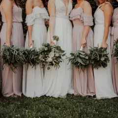 Un grupo de damas de honor en España, vestidas con trajes largos y fluidos en tonos pastel, sostienen ramos de eucalipto y flores blancas.