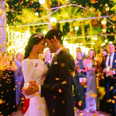Una pareja baila felizmente en su **noche de bodas**, rodeada de confeti dorado y luces cálidas. Invitados sonríen al fondo.