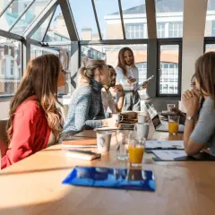 Un grupo diverso de profesionales participa en un workshop, discutiendo ideas alrededor de una mesa con laptops y bebidas.