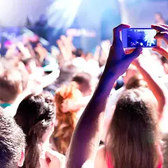 Manos sostienen un teléfono para grabar un evento comercial. La multitud levanta los brazos, disfrutando de la música y las luces.