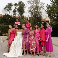 Grupo de mujeres sonriendo, vestidas con trajes rosas y morados. La novia, con un vestido blanco, está rodeada por su corte de honor, incluyendo a un hombre como dama de honor.