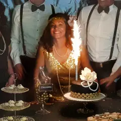Una mujer sonriente con un vestido dorado y perlas celebra en una fiesta temática de los años 20. Hay un pastel con bengalas y dulces.