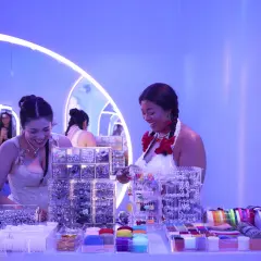 Dos mujeres sonríen en un puesto de accesorios para el cabello y cintas, parte de eventos de marca.