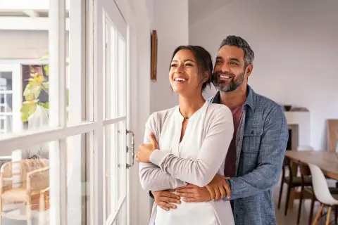 Pareja sonriente y abrazada, mirando por la ventana. Refleja cómo debe ser una relación de pareja: amor, apoyo y futuro compartido.