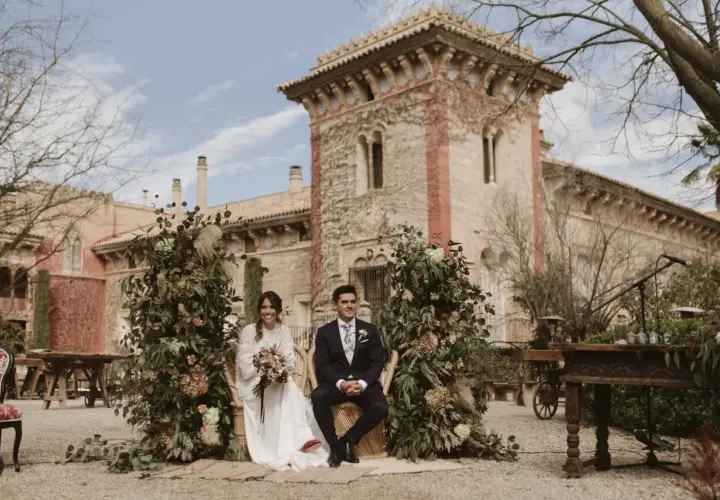 Novios sentados en sillas de mimbre, rodeados de exuberante vegetación y flores, con una arquitectura histórica de fondo. La decoración boda boho crea un ambiente romántico y natural.