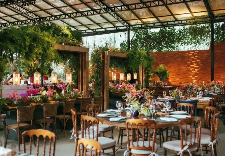 Salón de eventos decorado con abundante vegetación, flores y luces cálidas, con mesas puestas para una celebración.