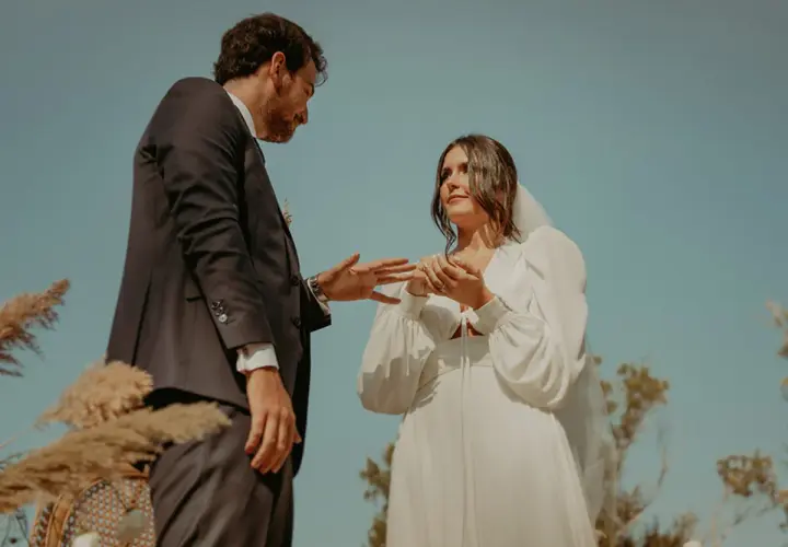 Novios intercambian anillos en una boda civil informal al aire libre. Un vestido blanco y un traje oscuro, rodeados de naturaleza.