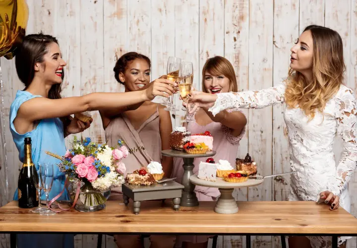 Cuatro mujeres brindan con copas de champán en unas fiestas familiares, rodeadas de pasteles y flores.