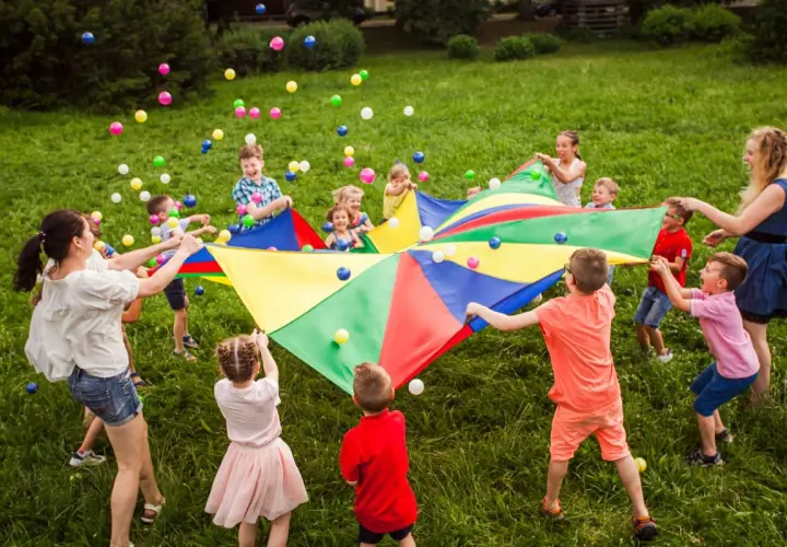 Niños y adultos juegan con un paracaídas de colores y pelotas, celebrando una fiesta de cumpleaños original en el césped.