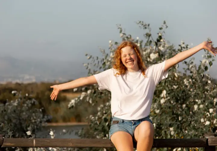 Chica pelirroja sonríe con brazos abiertos, disfrutando de la vida. Un símbolo de autoayuda y autoestima en armonía con la naturaleza.