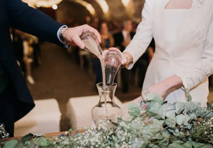 Manos de novios vierten arena en un jarrón, simbolizando su unión en una boda simbólica.
