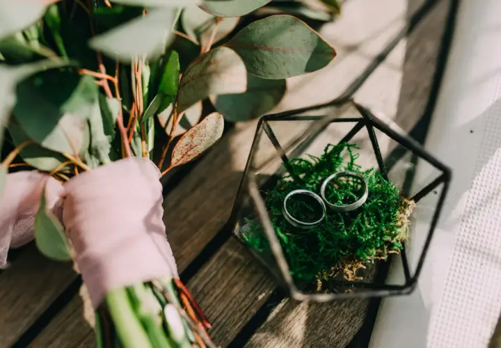 Anillos de boda en musgo verde dentro de un terrario de vidrio, junto a un ramo con tela rosa. Celebrando 5 años de casados.