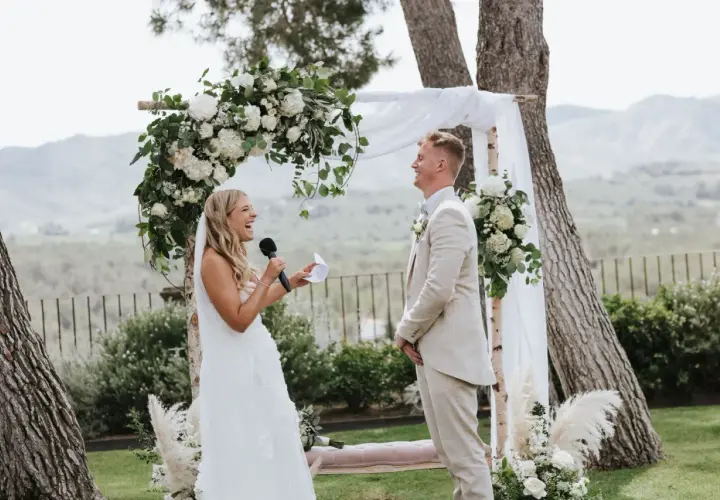 Novia ríe mientras lee sus votos, novio sonríe. Bodas sencillas pero bonitas, con arco floral y paisaje de fondo.
