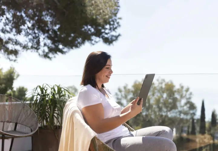 Mujer sonríe mientras trabaja en su tablet al aire libre, disfrutando de la vida de los trabajos para nómadas digitales.