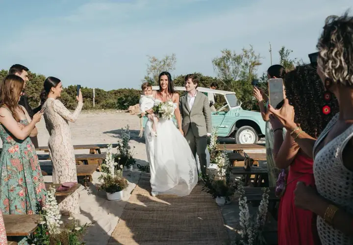 Novios caminan por la playa en una boda elegante. La novia lleva un niño en brazos. Un coche vintage espera.
