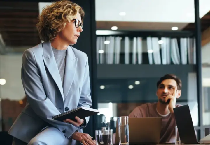 Mujer con gafas y traje gris, con cuaderno, en reunión. Podría ser parte de un curso organización de eventos gratis.