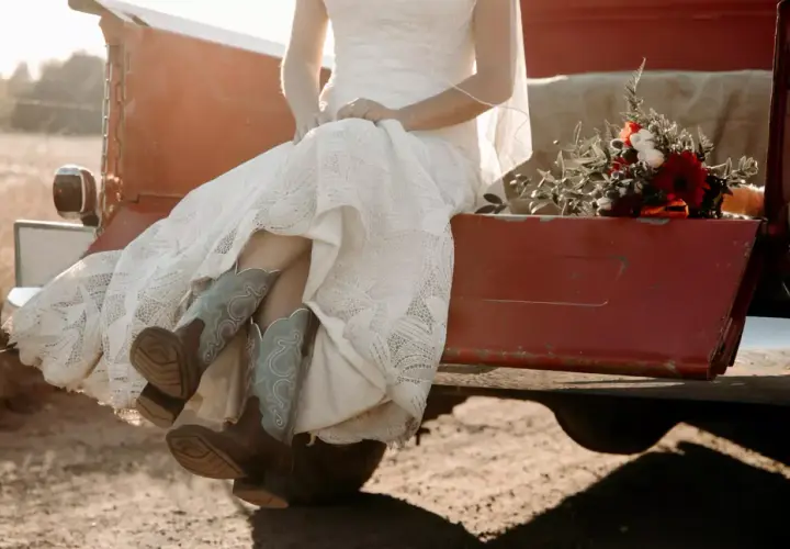 Novia con vestido de encaje y botas vaqueras, sentada en la parte trasera de una camioneta roja, con un ramo de flores. Un toque rústico para bodas vintage.
