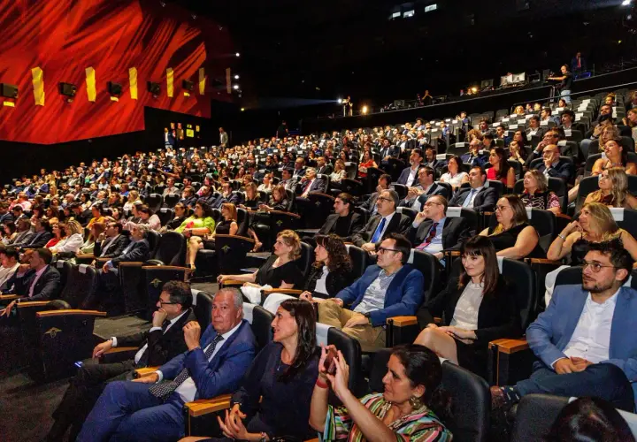Amplia audiencia en un auditorio oscuro, asistiendo a eventos de empresa. El telón rojo brillante y las luces del escenario iluminan la sala llena de gente.