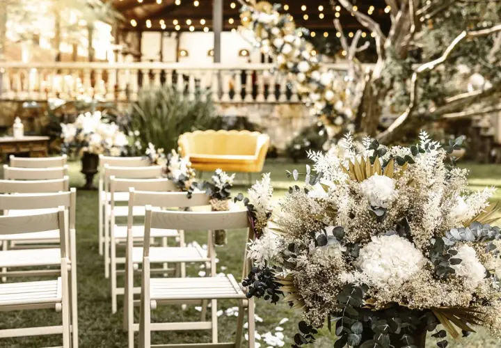 Un arreglo floral blanco y dorado decora un jardín para una boda civil sencilla y barata. Sillas de madera esperan a los invitados.