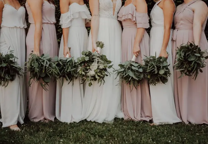 Un grupo de damas de honor en España, vestidas con trajes largos y fluidos en tonos pastel, sostienen ramos de eucalipto y flores blancas.