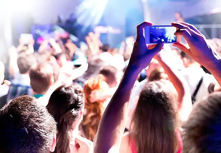 Manos sostienen un teléfono para grabar un evento comercial. La multitud levanta los brazos, disfrutando de la música y las luces.