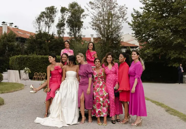 Grupo de mujeres sonriendo, vestidas con trajes rosas y morados. La novia, con un vestido blanco, está rodeada por su corte de honor, incluyendo a un hombre como dama de honor.