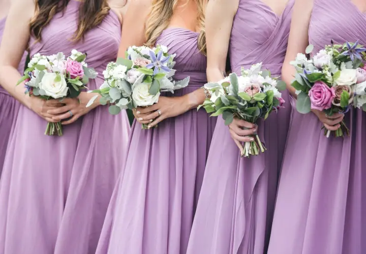 Damas de honor solteras, vestidas de lila, sostienen ramos de flores.