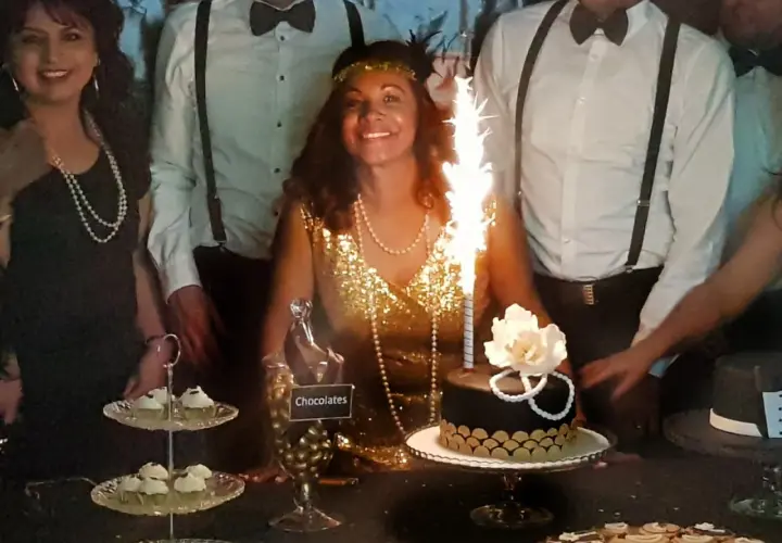 Una mujer sonriente con un vestido dorado y perlas celebra en una fiesta temática de los años 20. Hay un pastel con bengalas y dulces.