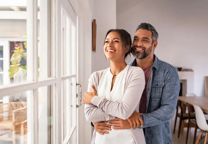Pareja sonriente y abrazada, mirando por la ventana. Refleja cómo debe ser una relación de pareja: amor, apoyo y futuro compartido.