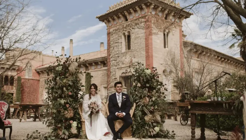 Novios sentados en sillas de mimbre, rodeados de exuberante vegetación y flores, con una arquitectura histórica de fondo. La decoración boda boho crea un ambiente romántico y natural.