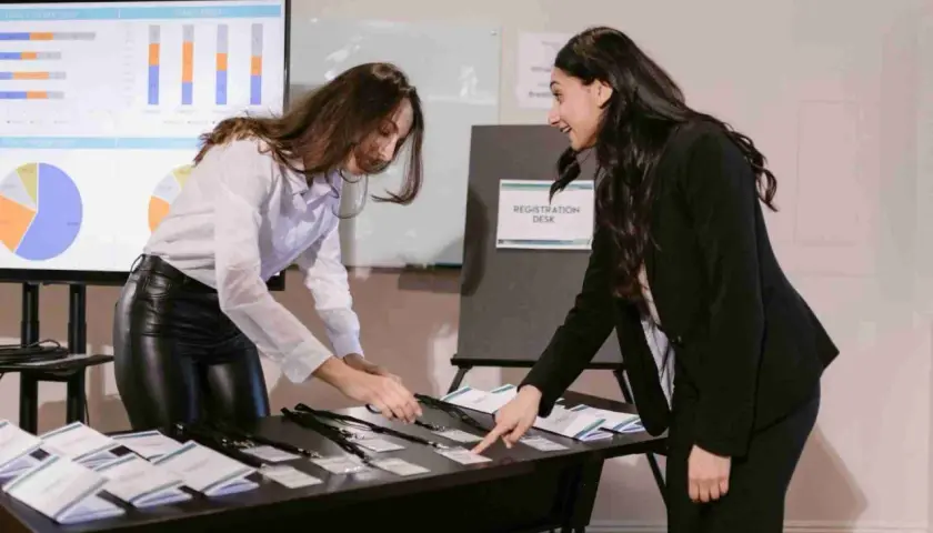 Dos mujeres organizan acreditaciones en un mostrador de registro, preparándose para lo que es una convención.
