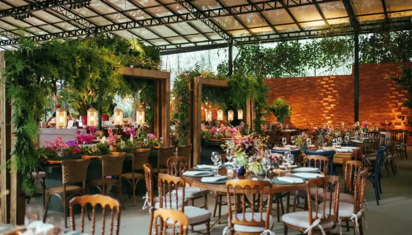Salón de eventos decorado con abundante vegetación, flores y luces cálidas, con mesas puestas para una celebración.