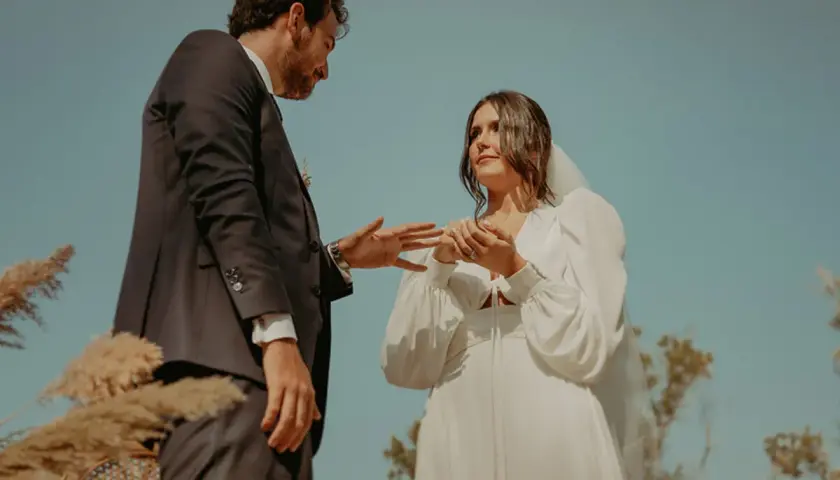 Novios intercambian anillos en una boda civil informal al aire libre. Un vestido blanco y un traje oscuro, rodeados de naturaleza.