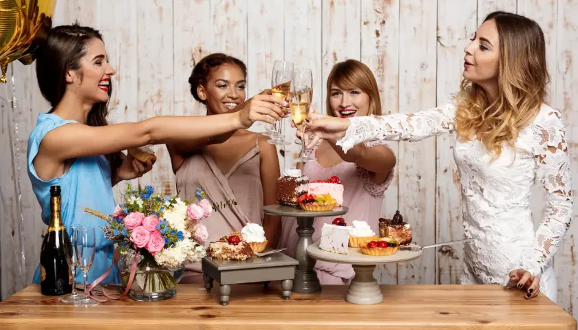 Cuatro mujeres brindan con copas de champán en unas fiestas familiares, rodeadas de pasteles y flores.