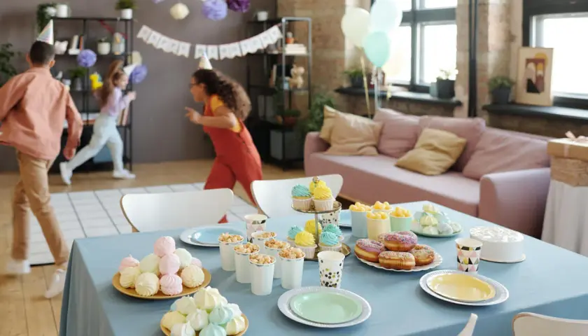Niños corriendo en una fiesta de cumpleaños, con mesa llena de dulces y pastel. ¡Ideas de qué hacer en una fiesta!