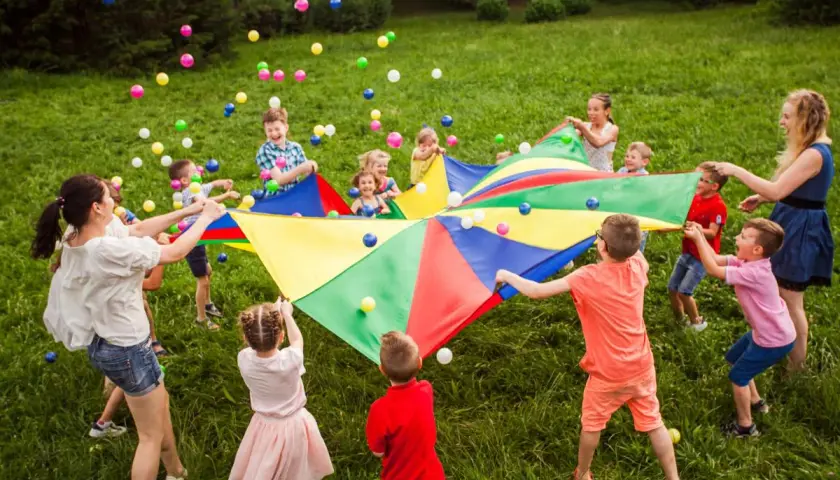 Niños y adultos juegan con un paracaídas de colores y pelotas, celebrando una fiesta de cumpleaños original en el césped.