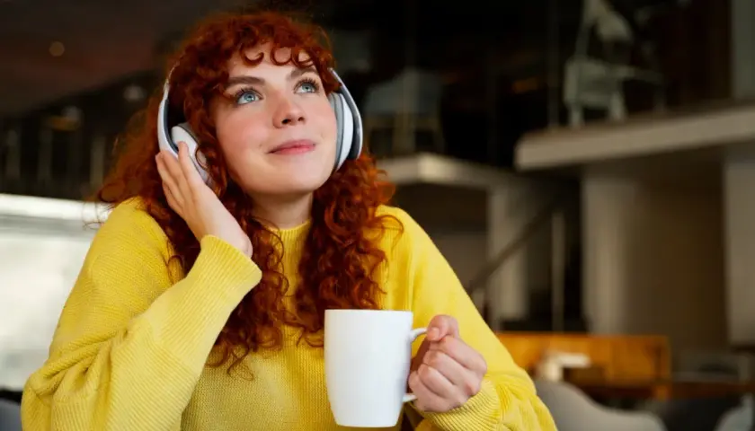 Chica con cabello rizado y suéter amarillo, con auriculares y taza, disfrutando de lo que parece ser la mejor manera de aprender inglés.