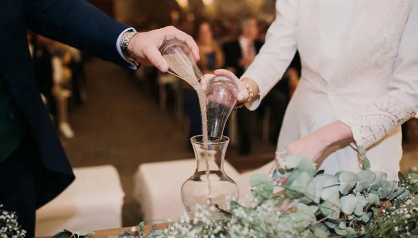 Manos de novios vierten arena en un jarrón, simbolizando su unión en una boda simbólica.